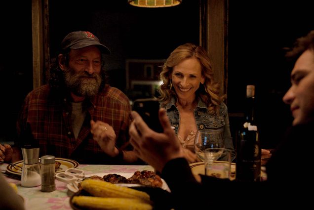 Photo of Emilia Jones, Troy Kotsur, Marlee Matlin and Daniel Durant in a scene from CODA. They are sitting around a table eating dinner. Kotsur wears a red sox baseball cap. Matlin smiles widely as Durant shows something to the family on his phone.