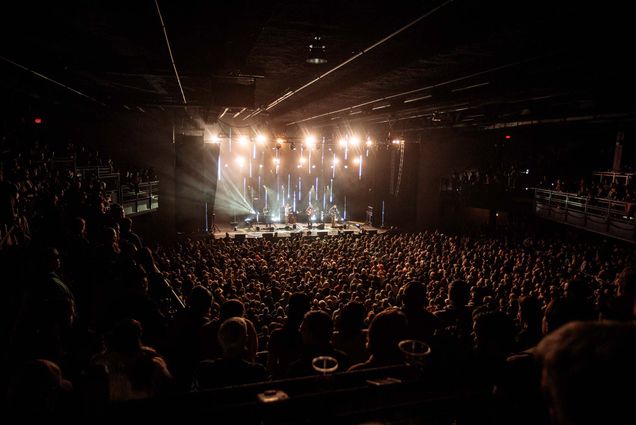 photo of the inside of Roadrunner during a concert with a full house. On stage, at back, a band is playing, lit by bright white lights. The photo is taken from above on the second floor balcony.