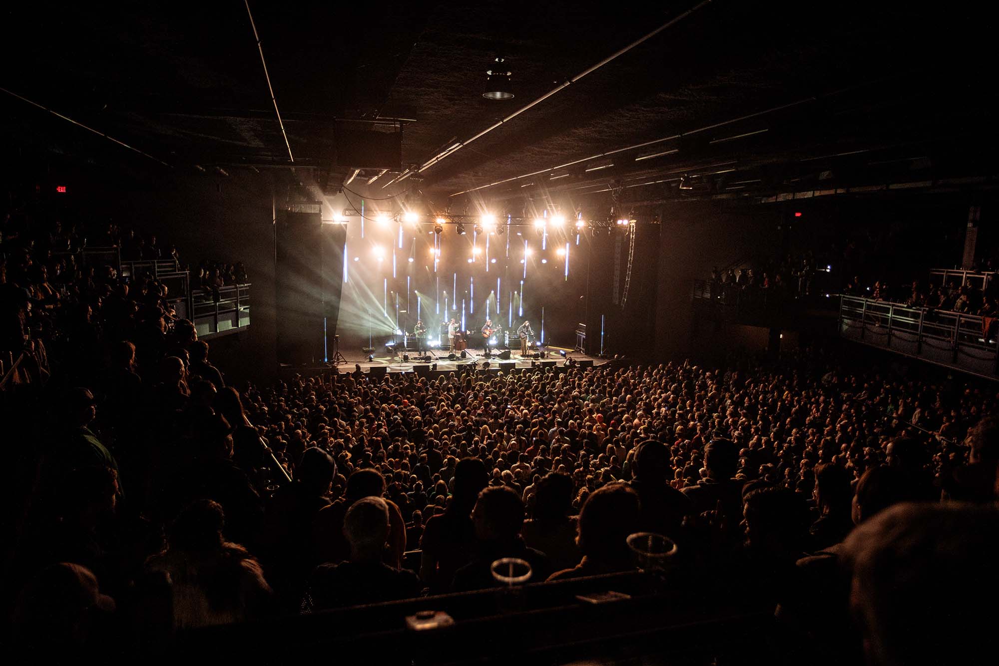 photo of the inside of Roadrunner during a concert with a full house. On stage, at back, a band is playing, lit by bright white lights. The photo is taken from above on the second floor balcony.
