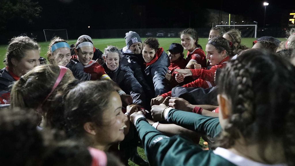 Photo of a women's soccer team huddle. Feldman is seen back left extending her arm to the center of the circle. The women smile, look excited, and are bundled up for an evening game late in the season.