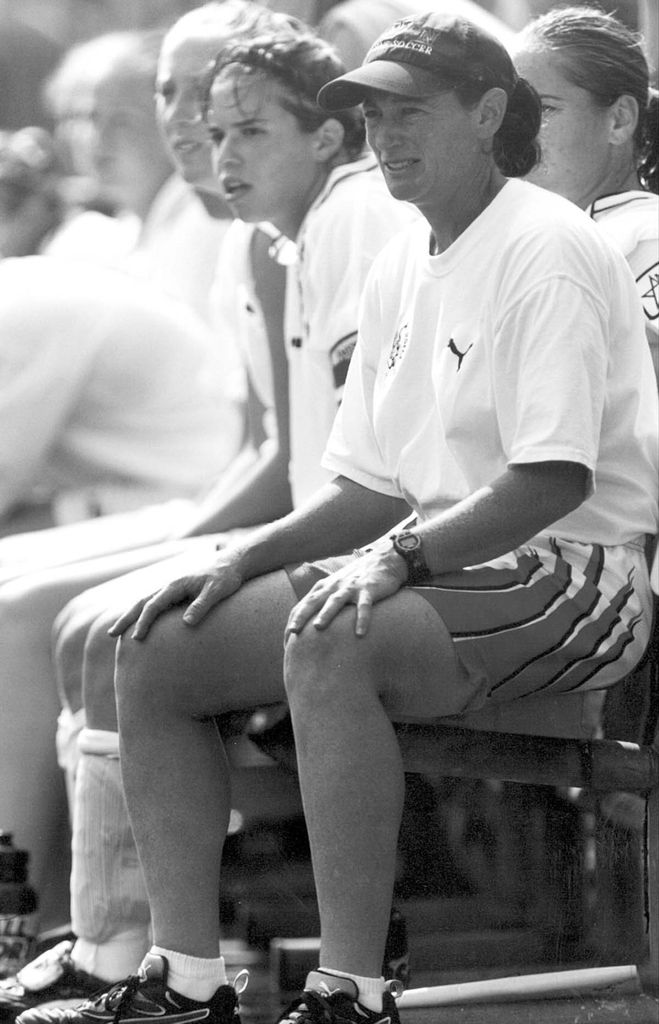 Black and white photo of Feldman, wearing a white shirt and red shorts with a white stripe and BU hat, sitting on the bench with women's soccer players.