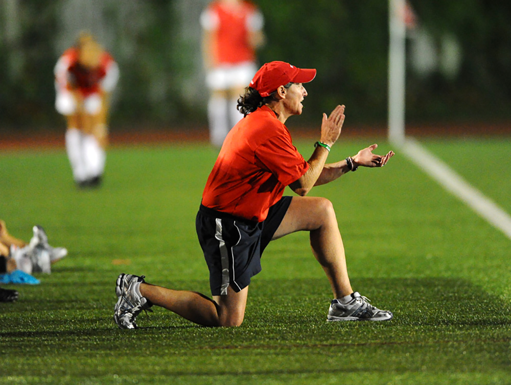 Photo of Nancy Feldman, a White middle-aged woman, kneeling along the side of a turf soccer field and clapping her hands during a soccer match. She wears a red hat, red shirt and dark gray shorts.