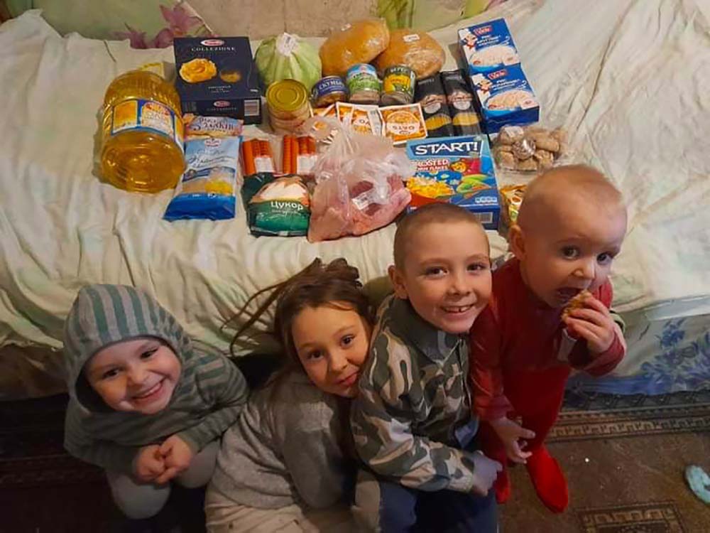 Photo of four small Ukrainian children, 3 boys and a girl, about 5 and under, in what looks to be their pajamas, posing in front of a bed filled with food. Food includes oil, pasta, onions, and other dry goods.