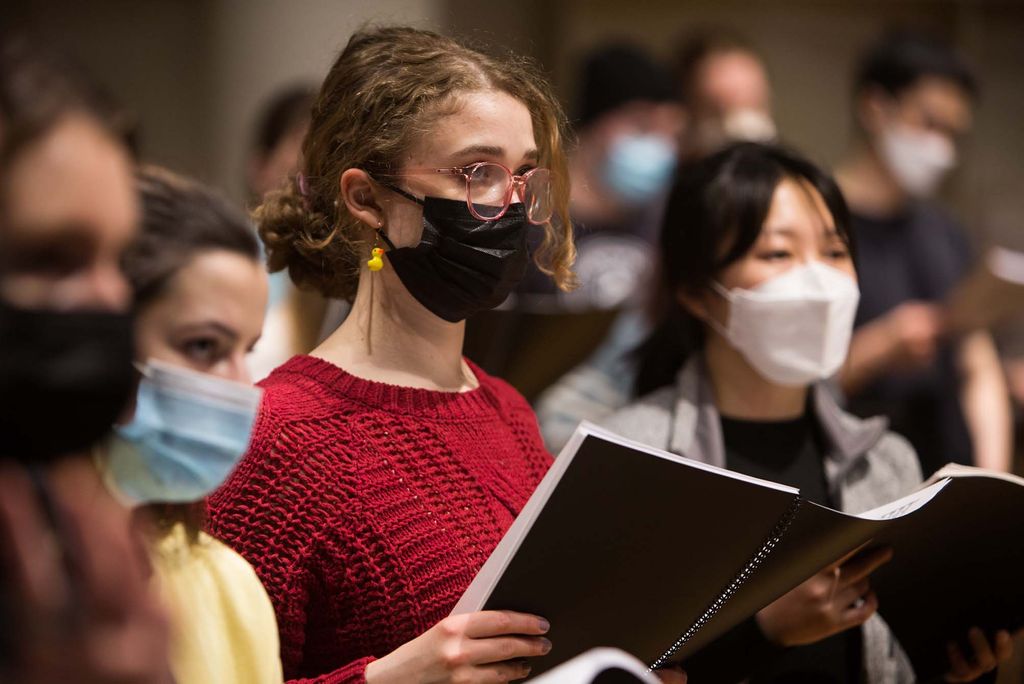 Photo of Hannah Voteur (CAS’22), a white woman with wavy dirty blonde hairy with a black face mask on, rehearsing with the Symphonic Chorus at the CFA Concert Hall. She wears a red sweater and other students are seen around her.