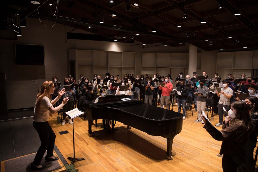 Photo of Dr. Mariah Wilson, Director of Choral Activities and Chair of the Conducting Department (left), directs the School of Music rehearsal on Wednesday, March 30, 2022 at the CFA Concert Hall. Students stand at right, on the other side of the grand piano, with masks on and librettos open.