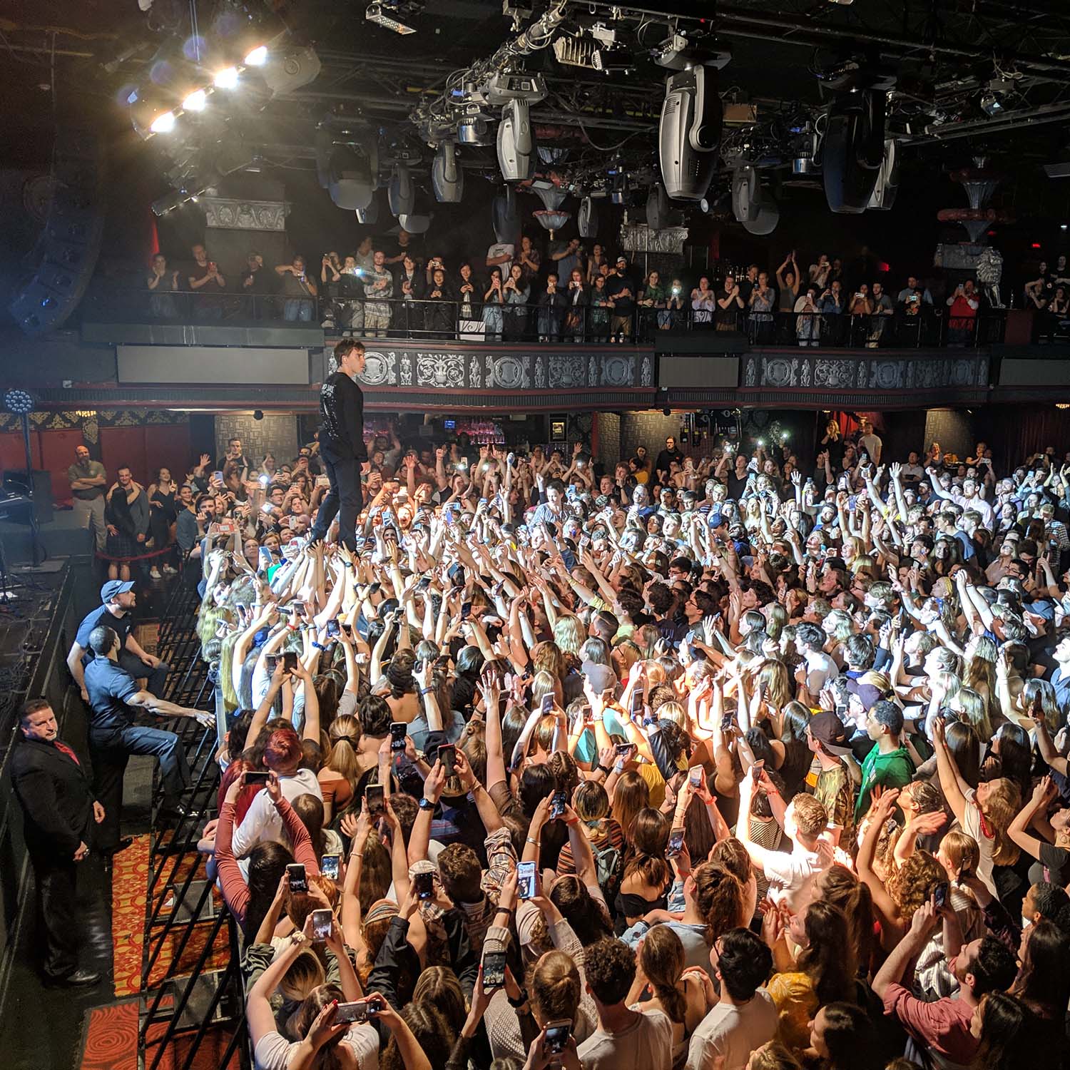 Photo taken during a Bad Suns concert at the House of Blues. The crowd has their arms raised and the lead singer is seen standing on top of some of the people in the first row by the stage. Folks are seen on the balcony as well.