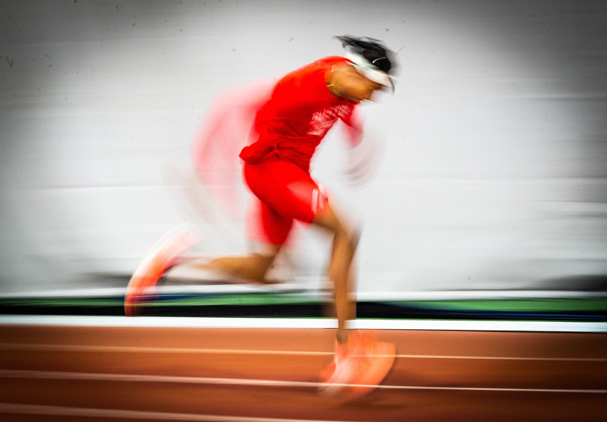 Photo of Toby Makoyawo mid run. A blurry image of a young, black man wearing head to toe red BU running gear is shown as he runs down in the BU Track and Tennis Center