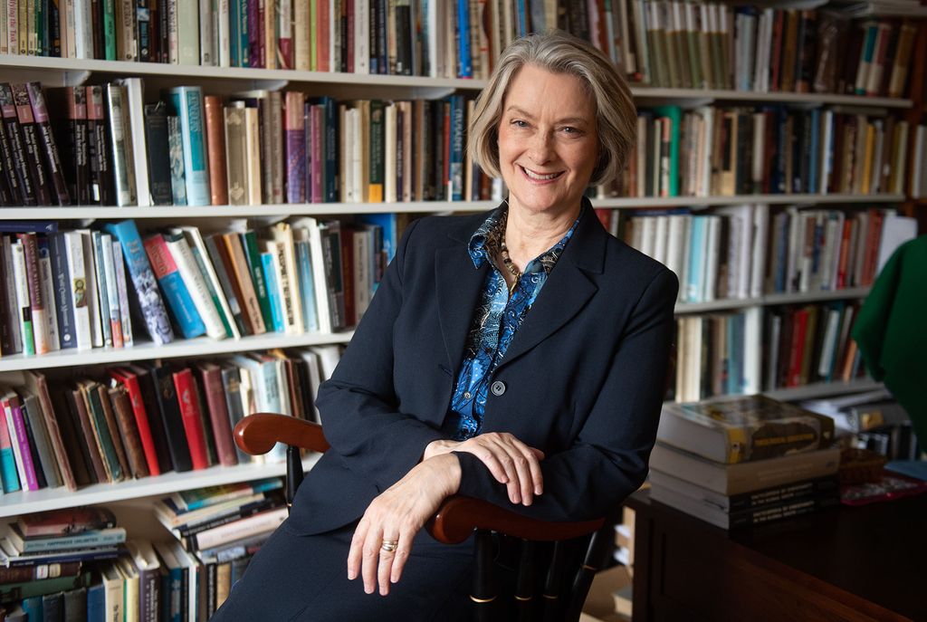 Photo of Professor Dana Robert of STH. She is a middle-aged White woman with short salt and pepper hair. She wears a black blazer and blue blouse, and smiles as she sits in a wooden chair, her hands crossed in her lap. Behind her, a large white bookshelf filled with books is seen.