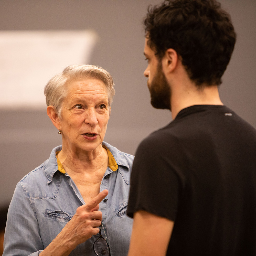 Photo of Judy Braha (CFA’08) speaking to a student during a rehearsal for Shakespeare in Love. Teh student has short brown hair and a beard, and wears a black t-shirt. Judy points at the student with her hand and appears to be giving directions.