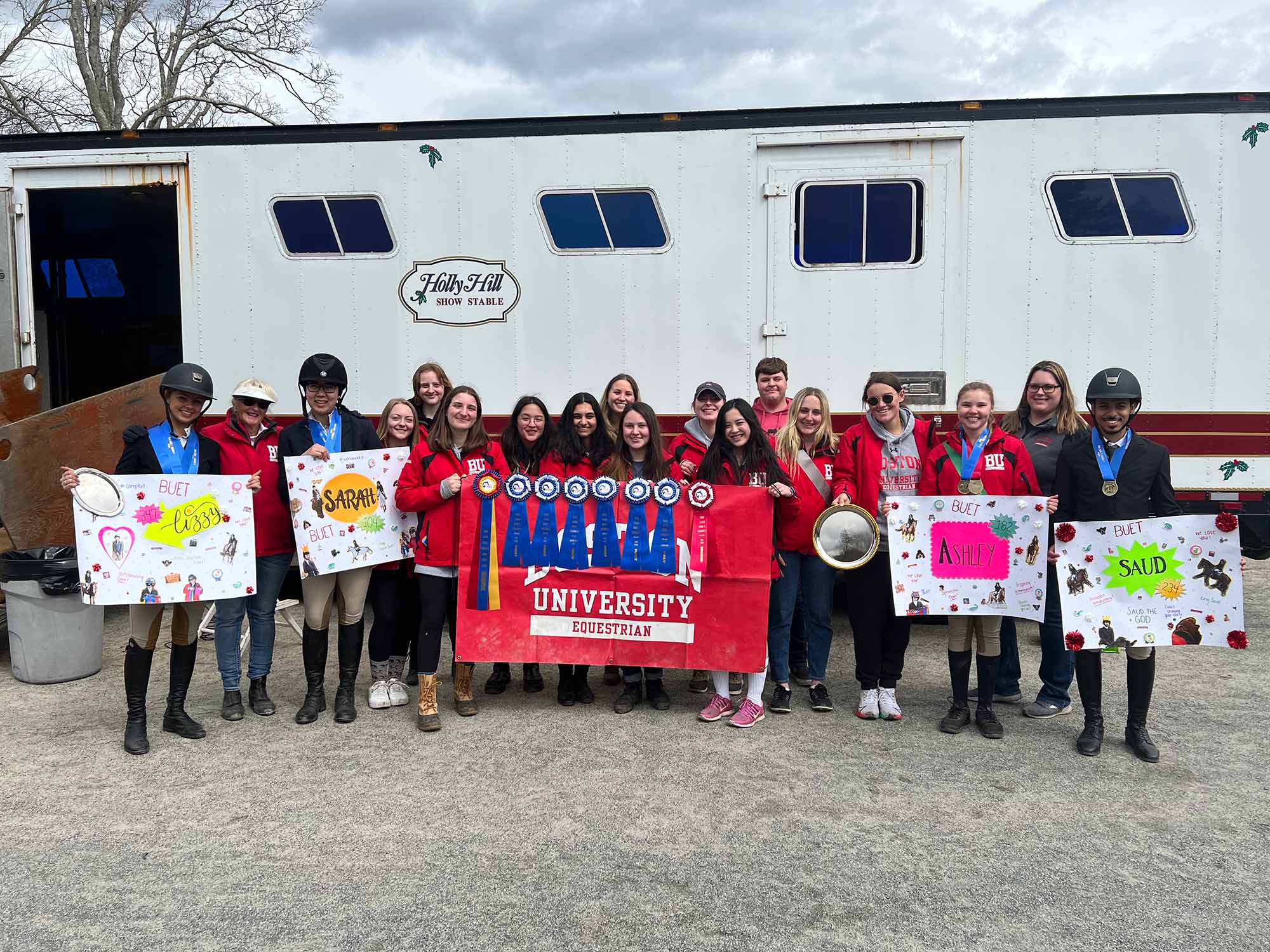 Photo of BU Equestrian Team members after a successful Regionals meet. A large group of young women and men stand in a row wearing a variety of red BU Equestrian jackets and black riding pose with signs and ribbon awards in front of a large white trailer with a label that reads "Holly Hill Show Stable" on the front.