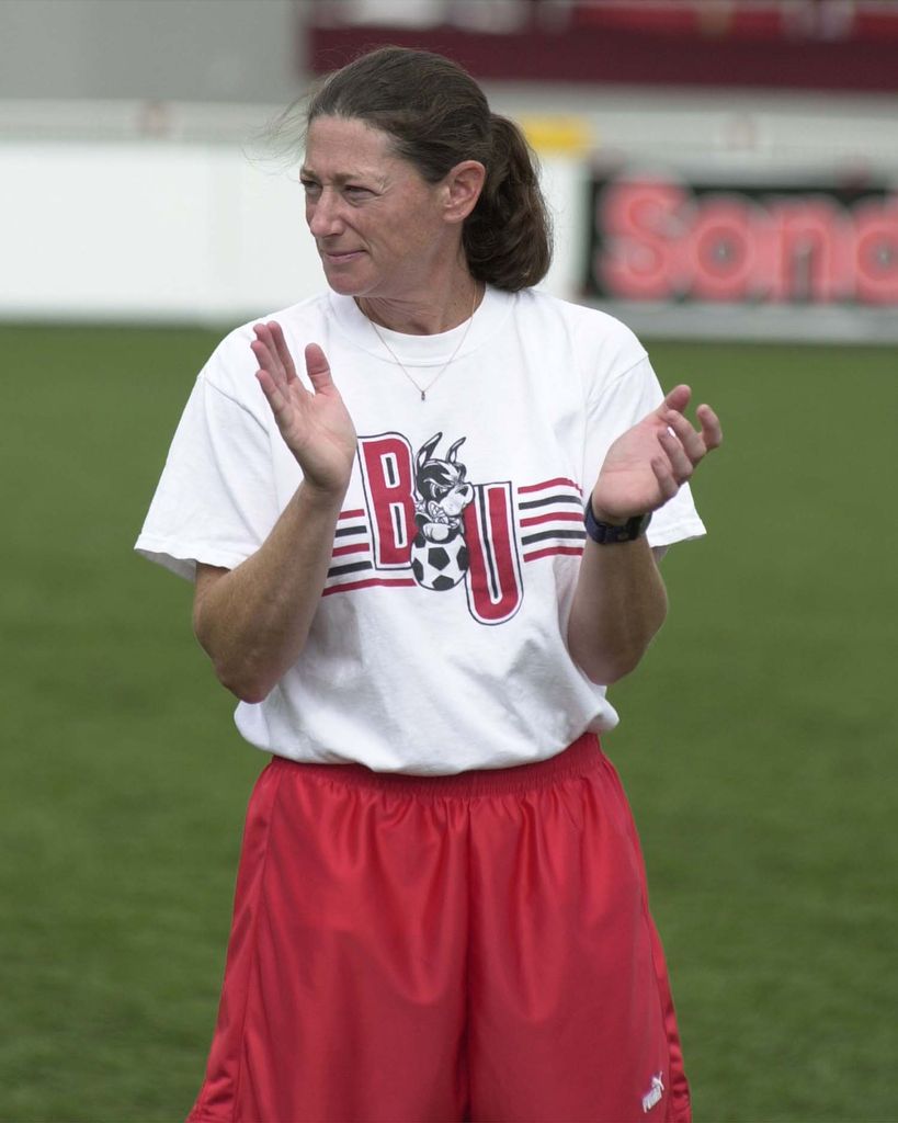 photo of Feldman, wearing red shorts and baggy white BU shirt with a terrier logo at center. She claps her hands, smiles slightly, and looks off to the side.