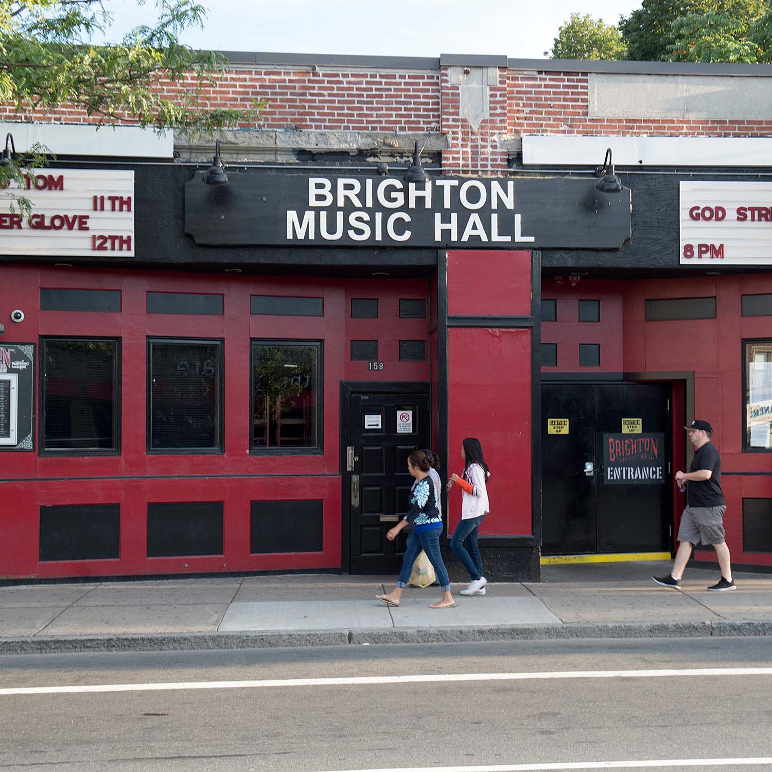 photo of the exterior of brighton music hall. Its facade is painted in red with blacked out windows. The sign over the door is black and reads "Bright Music Hall" in white. A few people walk by.