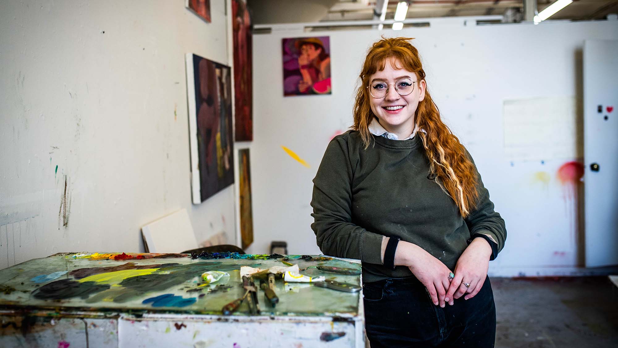 Photo of CFA graduate student, Eva Lewis, leaning against a white table covered with paint and paint mixing tools. She is a young white woman with long red hair and bangs, and wears round glasses and a green sweater. Behind her, other student's paintings hang on the wall.