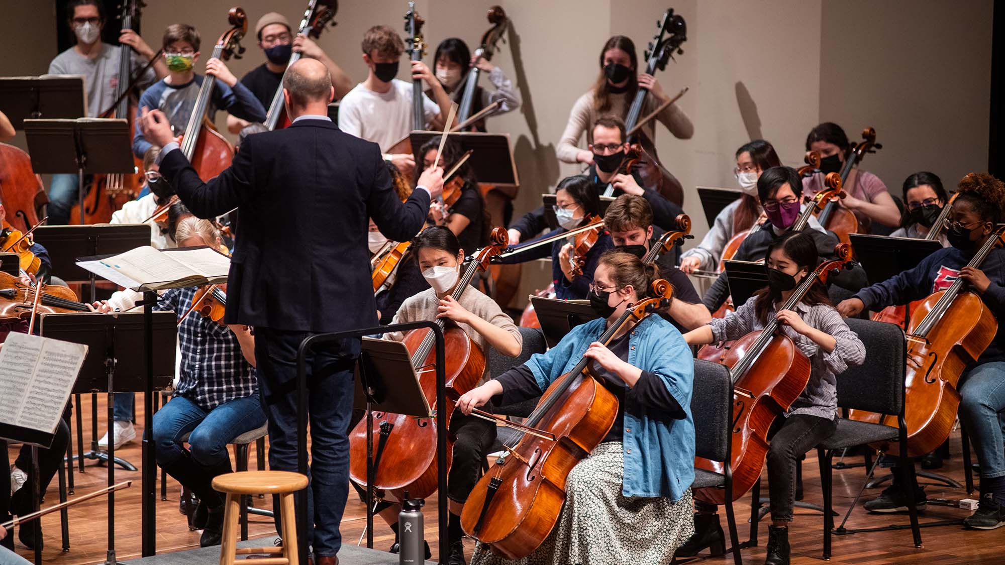 Photo taken from behind James Burton, Director of Orchestral Activities and Master Lecturer, Music during a BU orchestra practices at the Tsai Performance Center on March 30. Burton wears a suit and is in the middle of conducting; students wearing masks play string instruments at left.