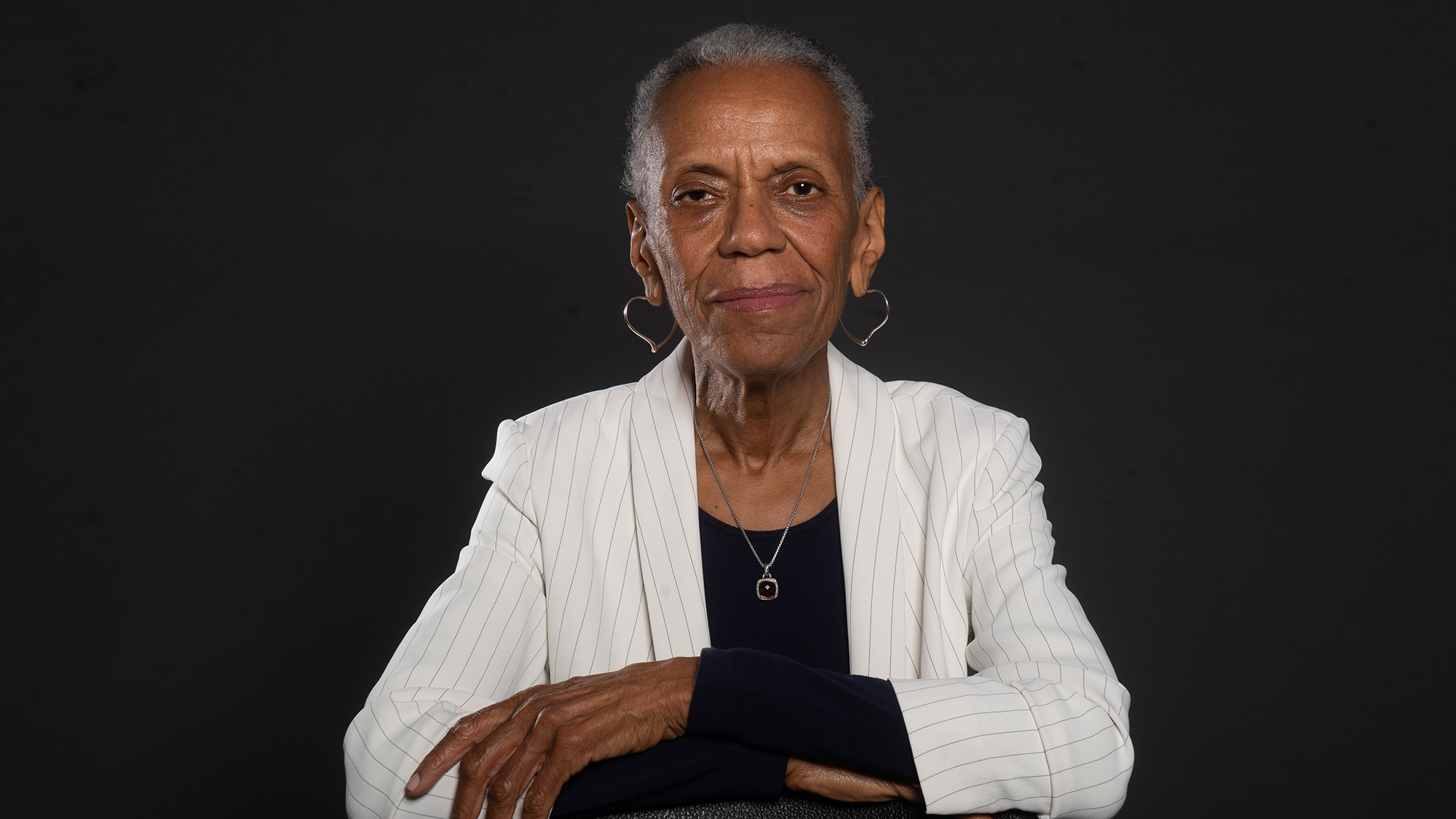 Studio portrait of Andrea Taylor. An older black woman with a short silver-grey hair and somber expression leans on a chair with arms crossed on top of the black chair. She wears a white blazer with light grey stripes, a long sleeved black shirt, and large heart-shaped earrings.