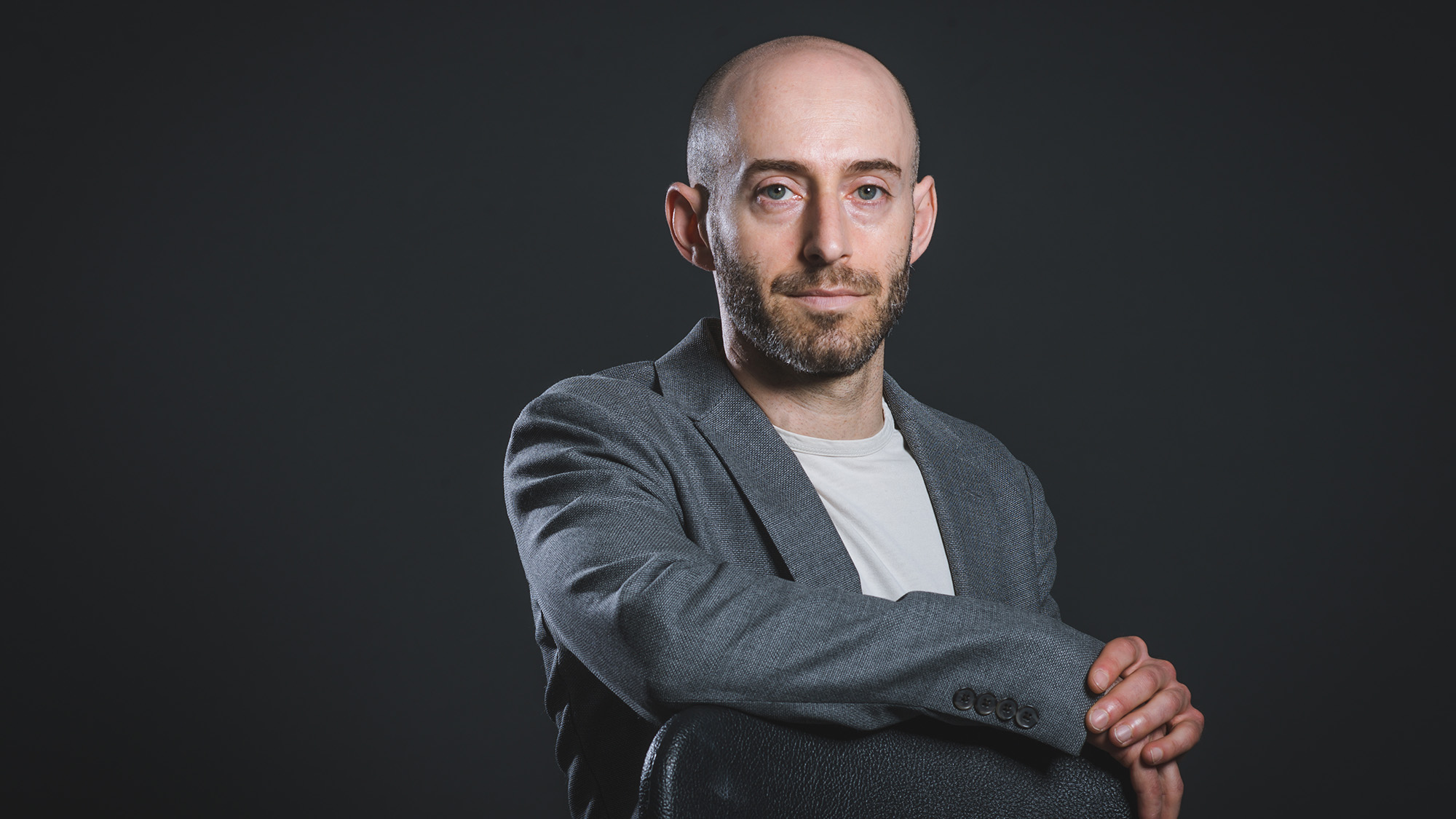 Studio portrait of Jonathan Feingold, Associate Professor of Law. A bald white man with a beard and mustache wears a somber expression and leans on a chair with one arm strewn across the top of the grey chair as the other hand holds it. He wears a grey blaze and white shirt.