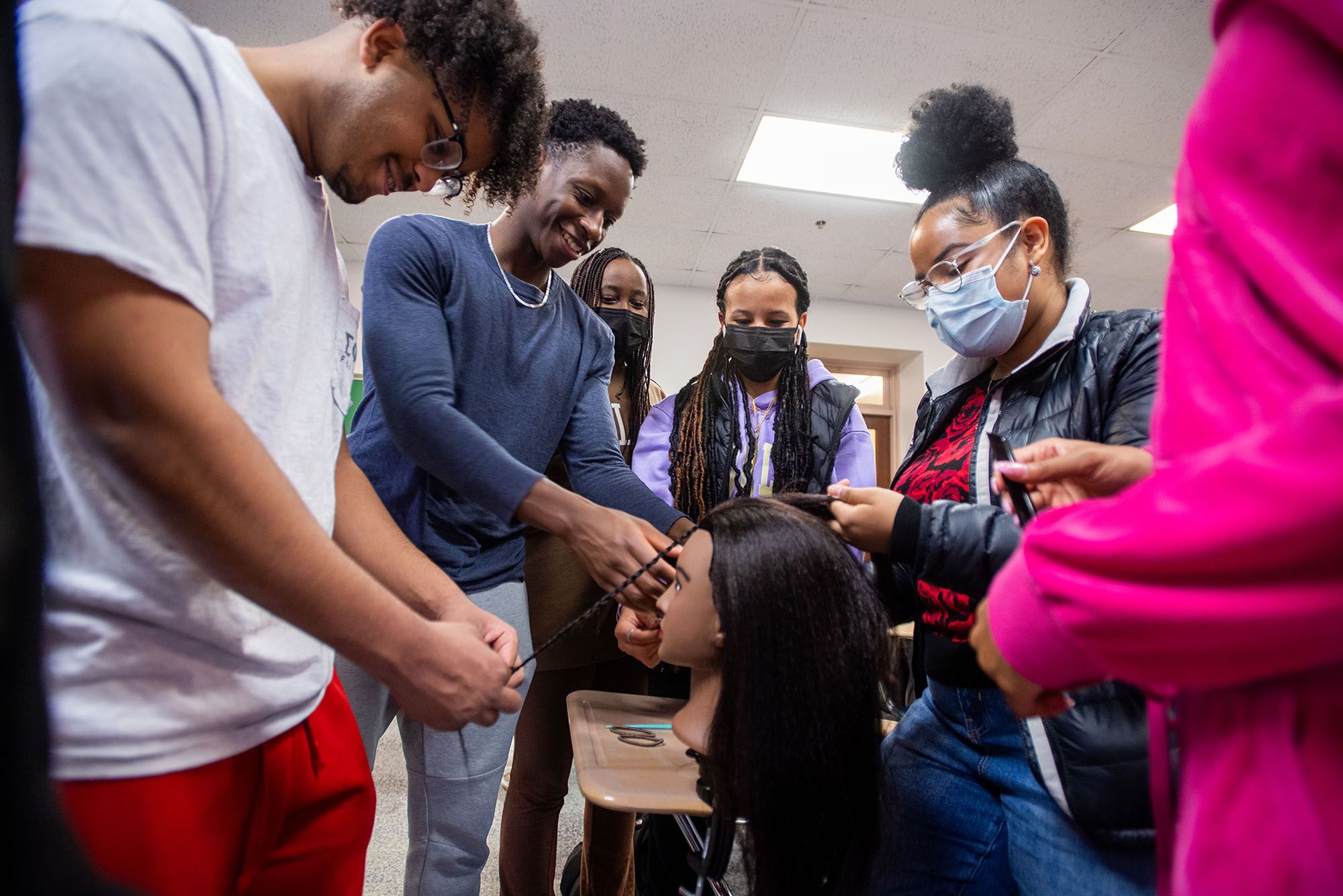 Photo of Aymen Yibrie, from left, N’Sikan Keendow, Abigail Mureithi, Axum Afework, Daniela Baez, and Naomi Boye work on their braiding during Naturally BU’s braiding demonstration at CAS. Various Black students stand around a mannequin head clipped to a desk as they practice and laugh. A few students wear masks.
