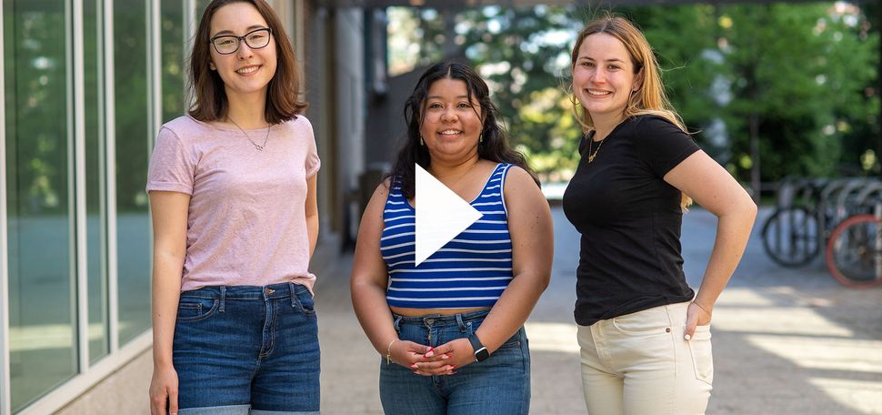 Photo of Newbury center grads Maelee Chen, from left, Cecilia Jarquin, and Annie Rose Hallett. Chen wears a pink tee shirt and jean short, Jarquin wears a striped blue and white tank top and blue jeans, and Hallett wears a black shirt and light tan pants. All 3 young women stand together and pose for the camera. A white triangle play button is overlaid in the middle of the photo.