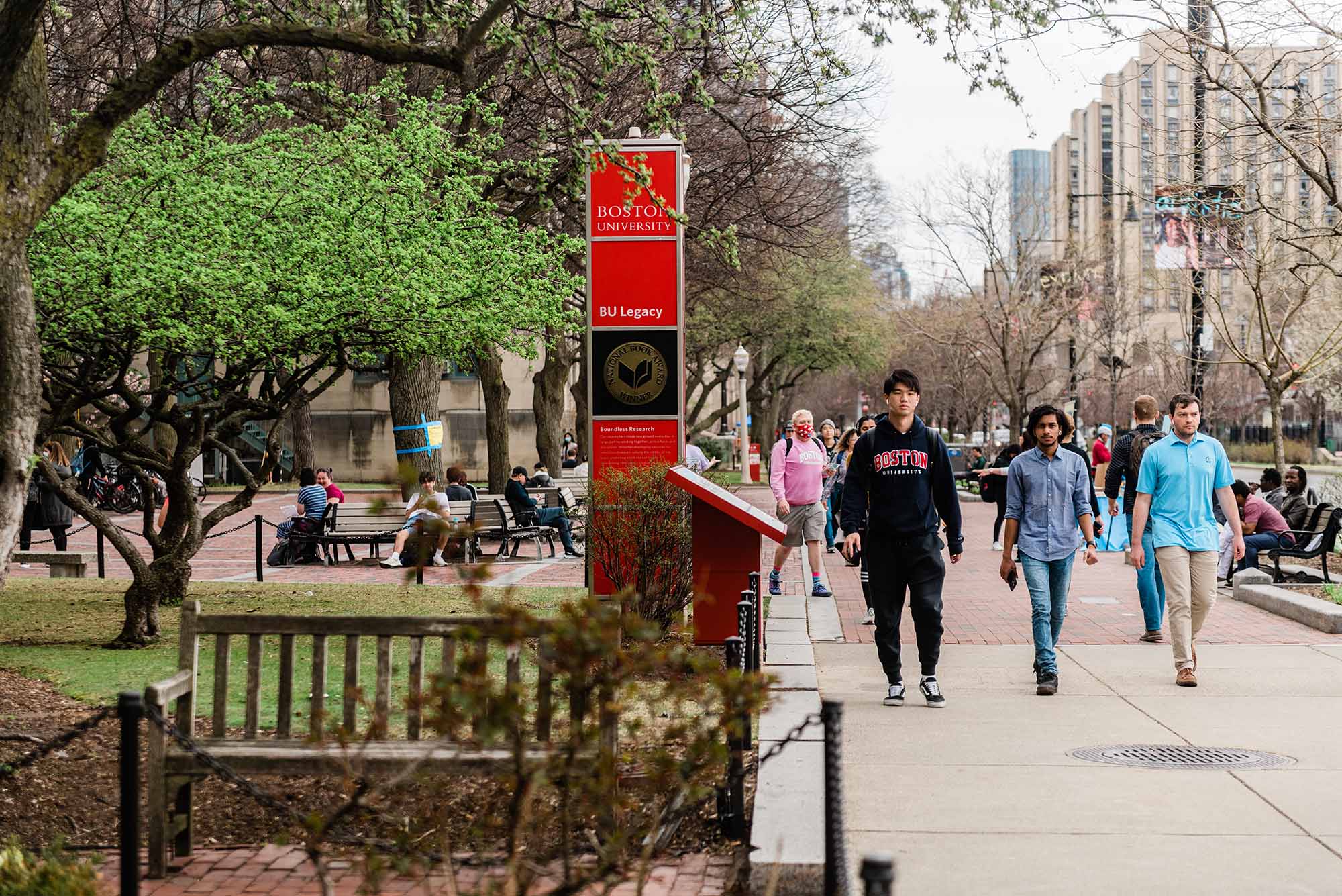 Photo of a diverse group of students walking down Comm Ave on a spring day. A red BU sign is seen at left, as well as a tree with budding green leaves.