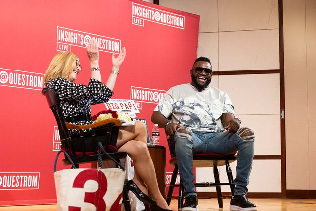 Photo of David Ortiz on stage with Questrom Prof. Susan Fournier in the Questrom auditorium during a one-hour Q&A on May 5. Fournier, a white woman who wears a black skirt and black and white blouse, claps her hands and smiles. Ortiz, a middle-aged Black man, is seen seated and smiling wide; he wears ripped jeans, a gray graphic tee and sunglasses. Next to Fournier on the ground is a canvas bag with the number 34, Ortiz's number, and behind them is a large red backdrop that reads "Insights@Questrom: LIVE"