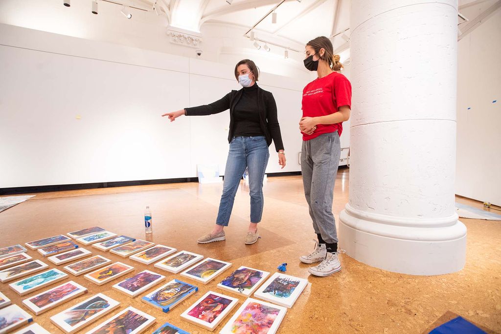 Photo of Leah Triplett Harrington, guest curator of this year’s BFA Thesis Art Exhibit, advising painting major Anna Frants (CFA’22) during installation of Frants’ work. Both wear face masks and stand looking at a series of paintings on the ground in front of them; each canvas is about 8x11 inches and features abstract portraits.