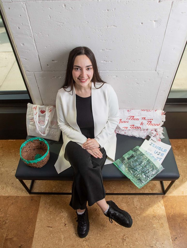 Photo of Kylie Carroll (CFA’22), a light-skinned woman with long brown hair who wears a white sweater, and black pants, sitting on a gray bench with her Single Use Society pieces at the BFA Thesis Art Exhibit at Stone Gallery May 5. The pieces include a woven basken to her right, and a few pieces made with what look like recycled white plastic bags.