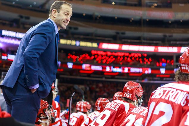 Photo of Jay Pandolfo, at left, in a blue suit, assistant coaching during a BU hockey game. The light of screens in the arena are seen behind him. At front right are the backs of BU Hockey players.