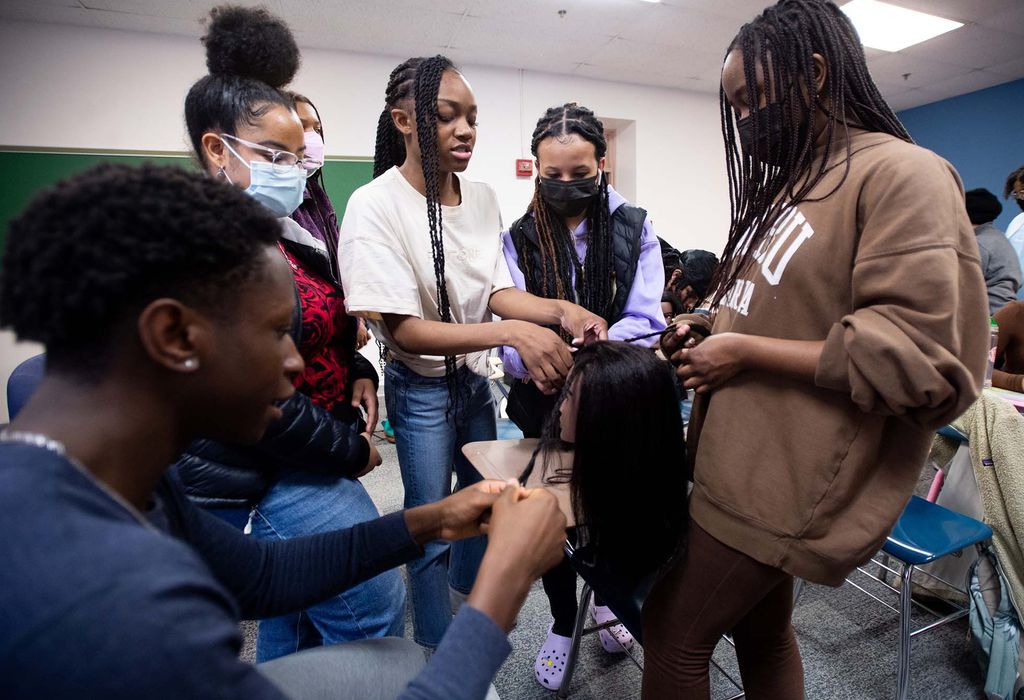 Photo of Tatiana Anderson, at center, teaching braiding to N’Sikan Keendow, from left, Daniela Baez, Axum Afework, and Abigail Mureithi during Naturally BU’s braiding demonstration at CAS. Various Black students stand around a mannequin head clipped to a desk as they practice and watch Tatiana braid on the head in the center. A few students wear masks.