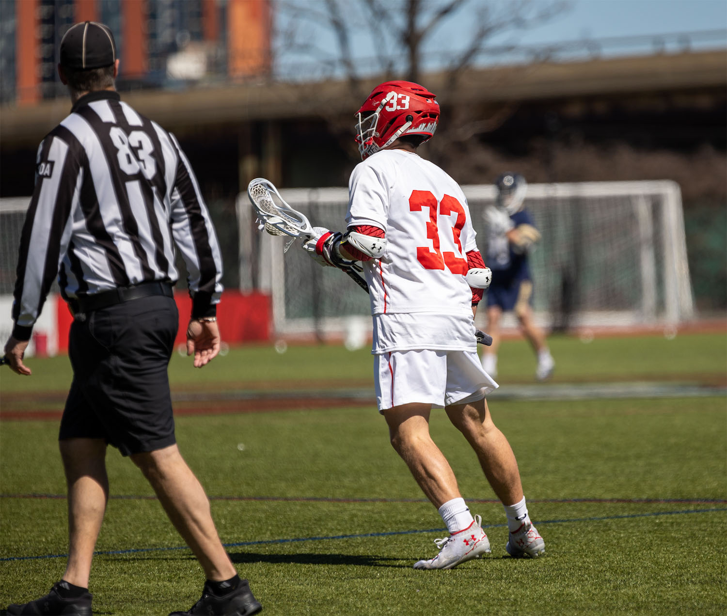 Photo of Matt Hilburn standing at the ready on Nickerson Field. He wears a white and red lacrosse uniform and red helmet and holds his lacrosse stick in front of him. A ref is caught standing next to him.