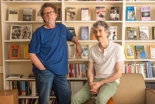Photo of Sven Birkerts, left, and William Pierce in the AGNI offices on Bay State Rd. Birkerts, a white man with light brown curly hair and a silver grey beard, wears a blue shirt and jeans as he leans back with one arm on a bookcase behind him. Pierce, a white man with grey and brown curly hair wearing a white short sleeved shirt and light green pants, sits on an armchair to the right of Birkerts, also in front of the bookcase. The bookcase behind spans the entire wall and is made of a light wood. Many books on it feature the title "AGNI"