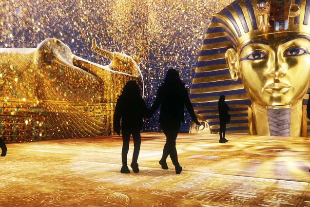 Photo of visitors stepping into an immersive gallery of golden treasure at the Beyond King Tut exhibit. Two people shown as shadows walk through a large golden exhibit. Large, golden King Tut head is seen at the entrance.