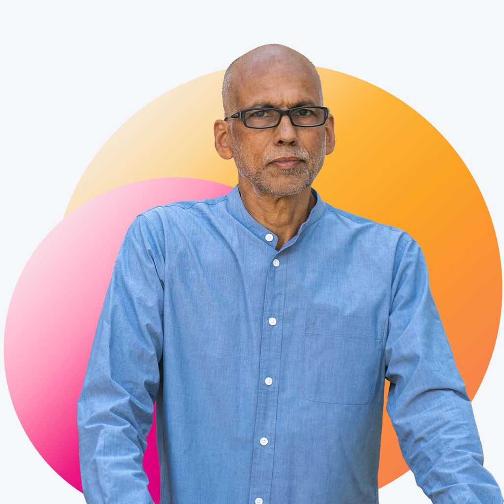 Portrait of Andre DeQuadros, a middle-aged man with brown skin, who wears a blue, collared shirt. He wears glasses and slightly smiles. Decorative pink and orange spheres are photoshopped behind his portrait.