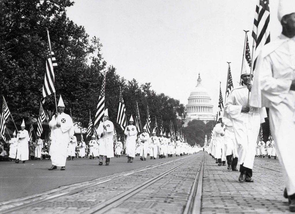 Black and white photo of a Ku Klux Klan on parade along Pennsylvania Avenue in Washington, D.C. on August 8, 1925. It was estimated that nearly 60,000 klansmen marched in the parade while nearly a million persons viewed the strange spectacle. The klansmen appear in their characteristic white hoods and pointed hats. They carry American flags, and march in long rows. The capital building is seen in the background.