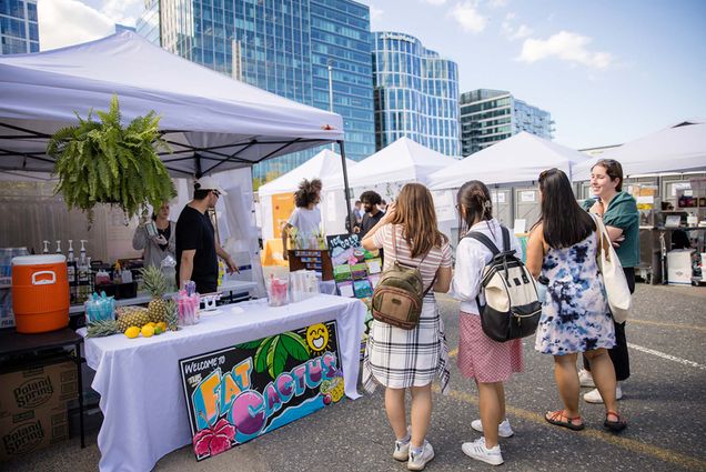photo of a group of young women with small backpacks and knee-length skirts or dresses stopping at a booth at the market called "fat cactus." The table underneath a white pop-up tent has fruits on it, such as pineapple, and a sign below the table reads "Fat Cactus" and is decorated with a smiling sun, a palm tree, and a flower. other white tents are seen in the background. A man in a white hat and black shirt, as well as a woman in gray sweatshirt that holds up a bottle of something are seen behind the table.