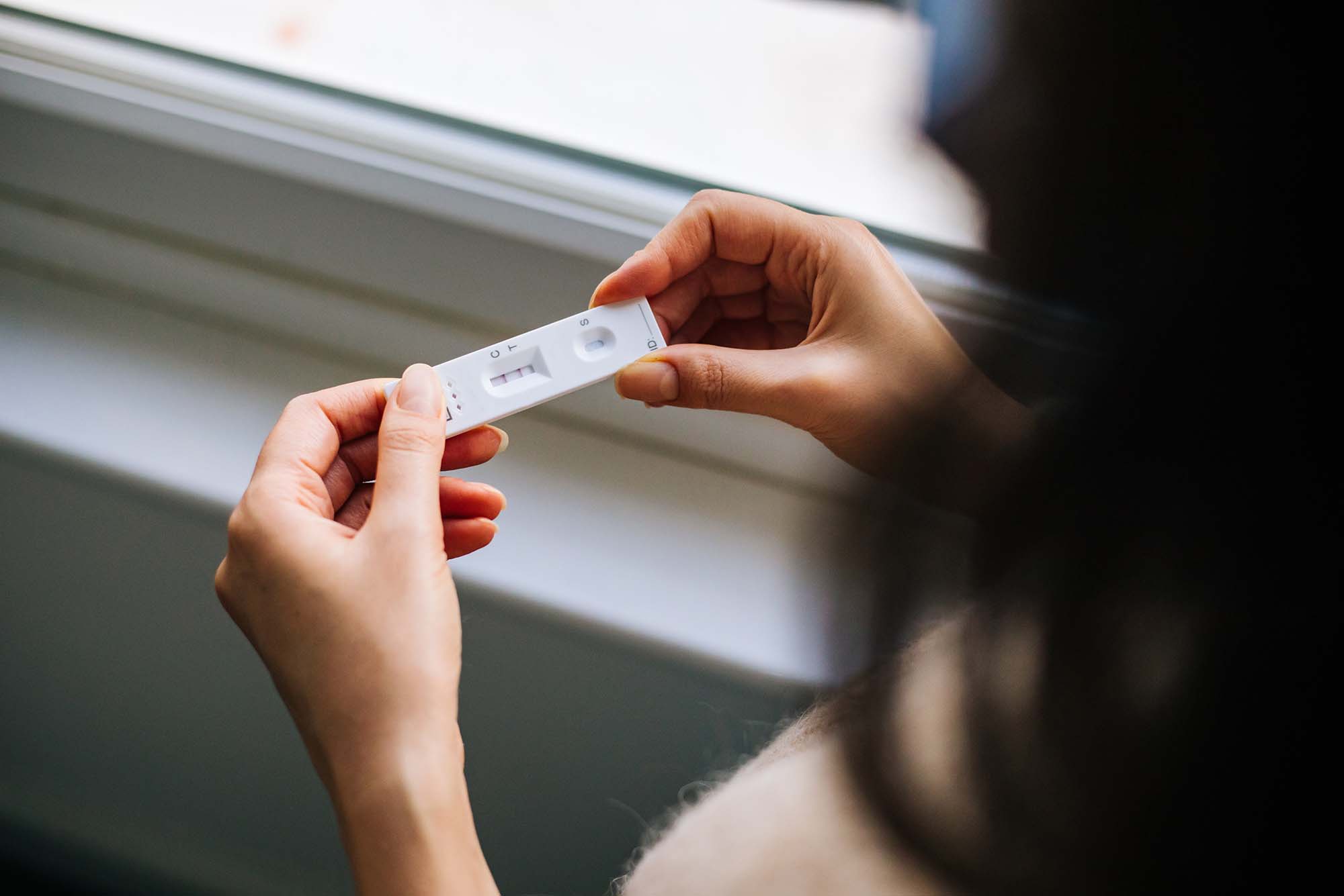 Photo taken from behind as White woman holds a Covid-19 Rapid At home test in her two hands. The back of the women's head is blurred. The test is a small white piece of plastic with lines on it.