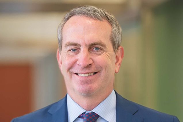 Photo of Anthony Hollenberg, a middle-aged White man with short gray hair. He smiles, wears a blue suit with a dark blue tie with red spots, and poses on a blurred background, which looks like the inside of a building.