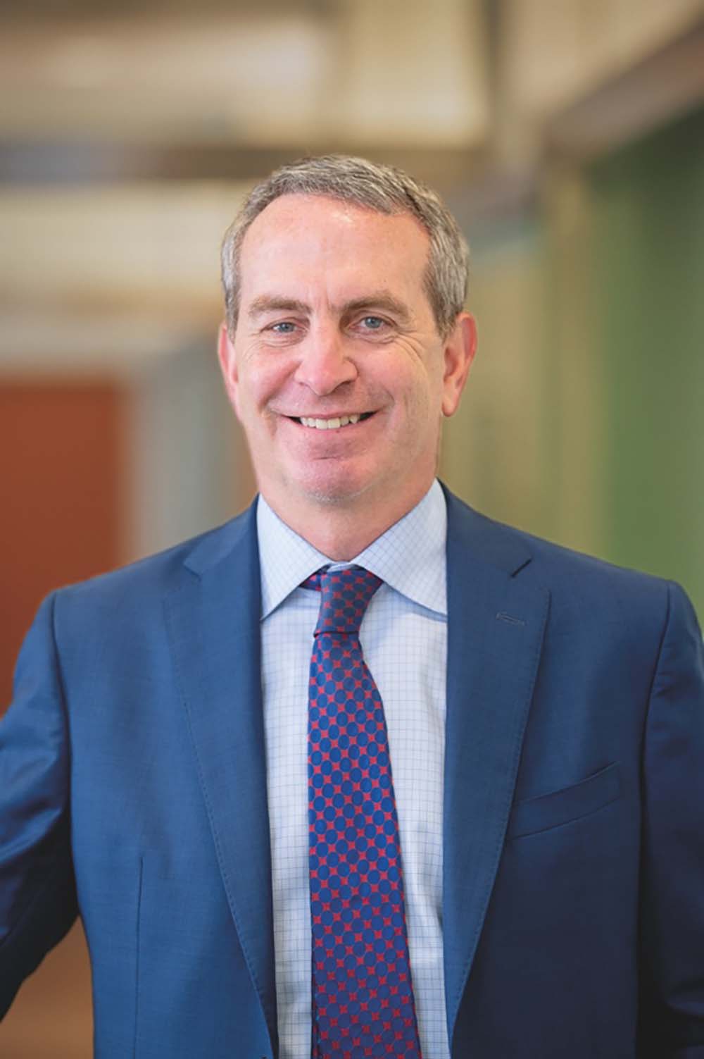 Photo of Anthony Hollenberg, a middle-aged White man with short gray hair. He smiles, wears a blue suit with a dark blue tie with red spots, and poses on a blurred background, which looks like the inside of a building.