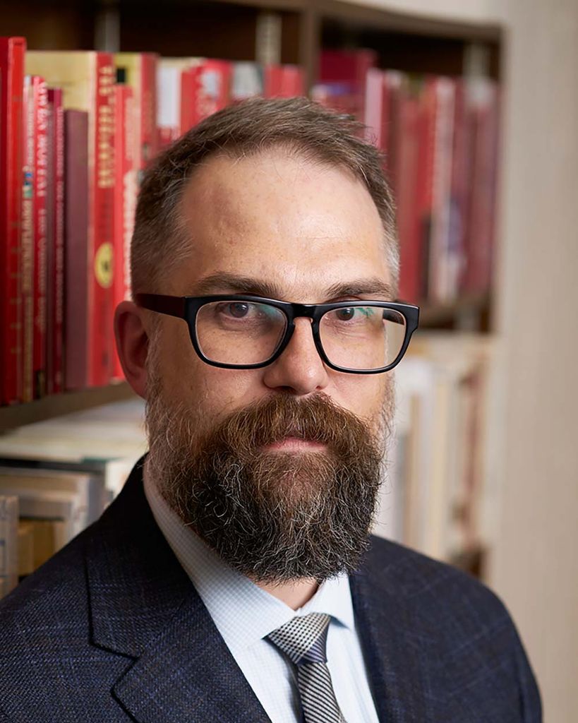 Photo of Mark Newton, a youngish White man with short brown hair, a long, brown beard and dark-rimmed glasses. He wears a suit and tie and stands in front of book case and makes a serious face.