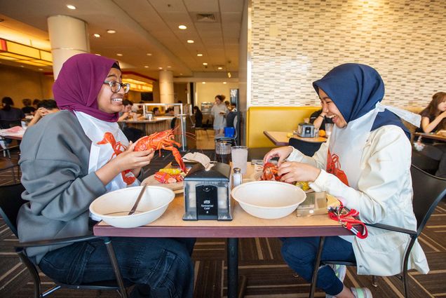 Photo: Rena Nurhussien (CAS’26), left, and Amira Badruddin (ENG’26) enjoy their special meal during Lobster Night at Warren Towers. A young woman on the left wearing a burgundy head scarf holds up a lobster excitedly as a young woman on the right wearing a navy headscarf laughs and holds her own lobster.