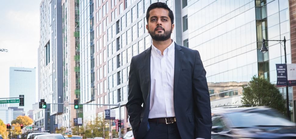 Photo: SHA faculty member Makarand Mody poses for a photo on Boylston Street in Boston. Modt poses in a suit ensemble in front of a city scene of high rise buildings and busy streets.