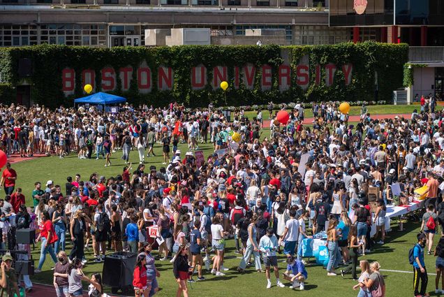 Photo of a crowd of BU students checking out booths, hanging around, eating, and just generally having fun on Nickerson Field, a soccer field with a track a red track surrounding it. The brick walls on one side of the field have large red letters that read "Boston University"