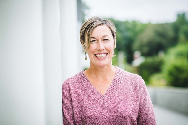 Photo of Kara Cattani. A white woman with short blonde hair leans against a white pillar and smiles for the camera. She wears a pink sweater and black pants.