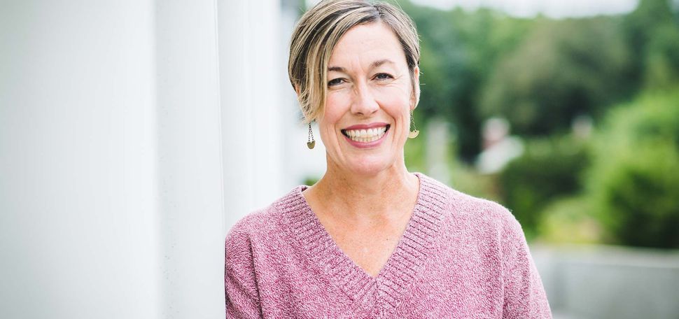 Photo of Kara Cattani. A white woman with short blonde hair leans against a white pillar and smiles for the camera. She wears a pink sweater and black pants.