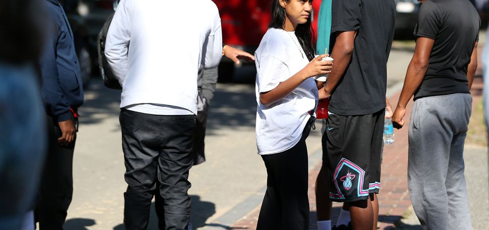 Photo: A group of migrants huddle on a sidewalk in front of St. Andrews Episcopal Church. A young Latina woman wearing a large white shirt holds a white cup and looks off as other Latinx individuals stand and look off around ehr.