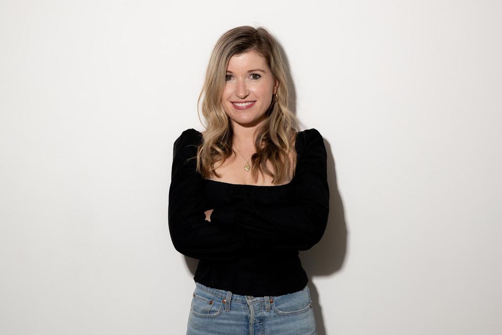 Photo: Studio headshot of Sarah Moret. A young white woman wearing a long-sleeved black shirt and jeans smiles in front of a white studio backdrop.
