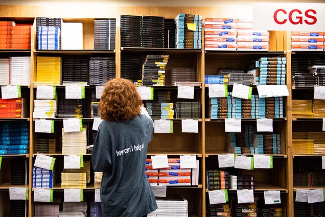 Photo of an employee of the BU bookstore taken from behind. They have short red, wavy hair and wear a large gray shirt that reads "how can i help" in white text. They pull a book from the shelf, which is about eight feet high and has several compartments, presumably one for each class. The stacks of books are labelled with white labels.