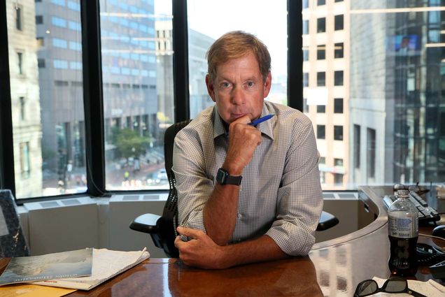 Photo of Brian McGrory, in his office at the Boston Globe. McGrory is a middle-aged White man with short blondish hair. He poses with his hand on his chin and holds a blue pen. He has a large desk that curves around to the back and his office has a large window that looks out onto downtown Boston. Papers and diet pepsi are seen in the foreground.