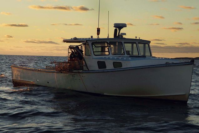 Photo of a small white lobster boat at sunset or sunrise. The sky is mostly yellow with some blue and a few thin clouds. On the boat, you can see what looks like two people working with lobster traps. Land is seen at right in the distance.