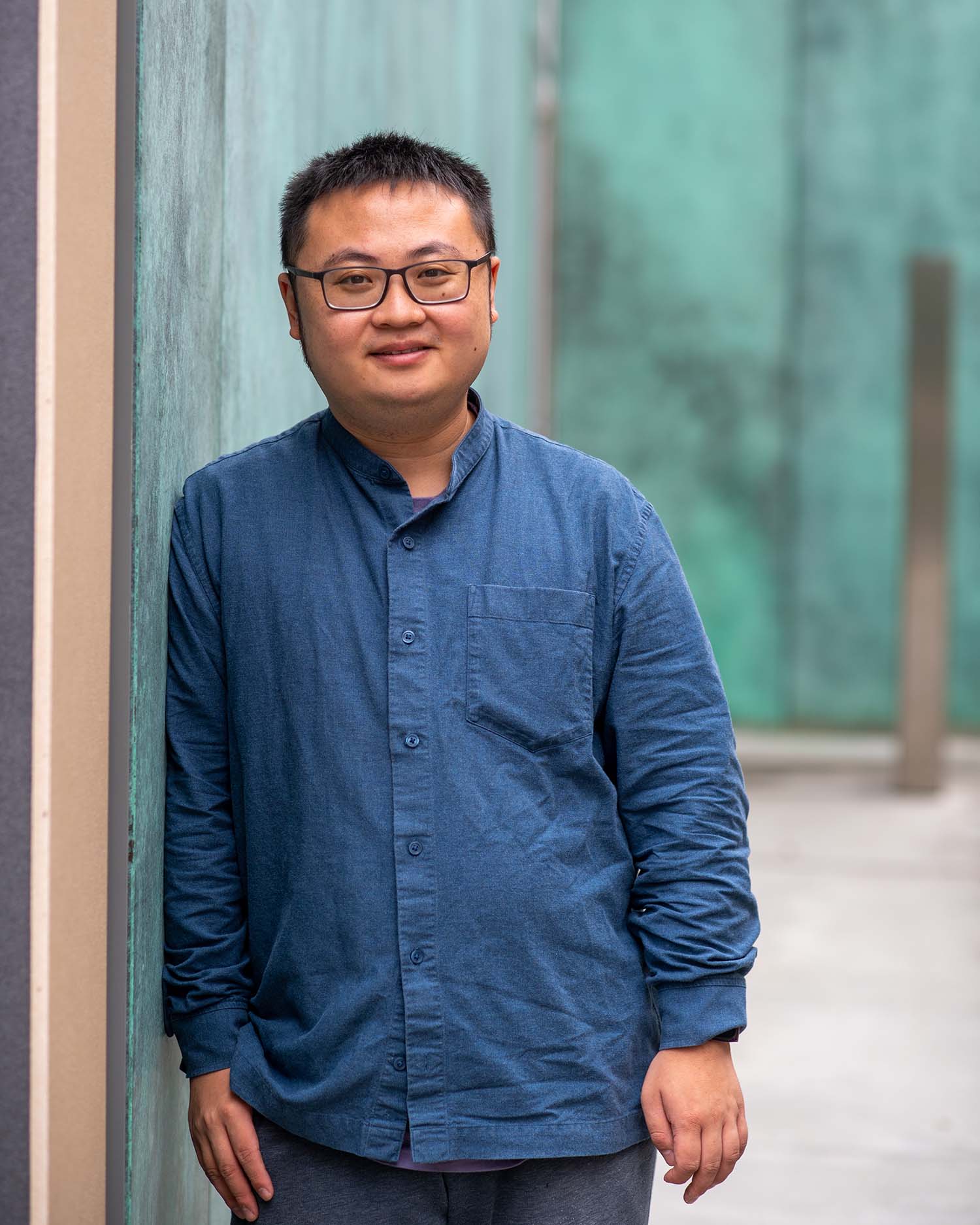 Photo of Yuandi Tang (COM’23), an international student from China, who wears glasses with black frames. He wears a blue button-down shirt, smiles, and leans against the exterior of building with rusted steel on the outside.