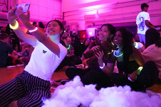 Photo of three young women of color taking a selfie during the Neon event during weeks of welcome. At left, Michelle Tian (COM'23), wears a white shirt, striped pants, and glowstick bracelets and holds out her phone to take the selfie. Her friends Jessica Lam (COM'24) and Anjali Zafiia (COM'23), to the right, sit behind her. One of the young women holds a teddy bear. A fluffy white substance is seen in the foreground; other students are seen sitting on the ground in the background.
